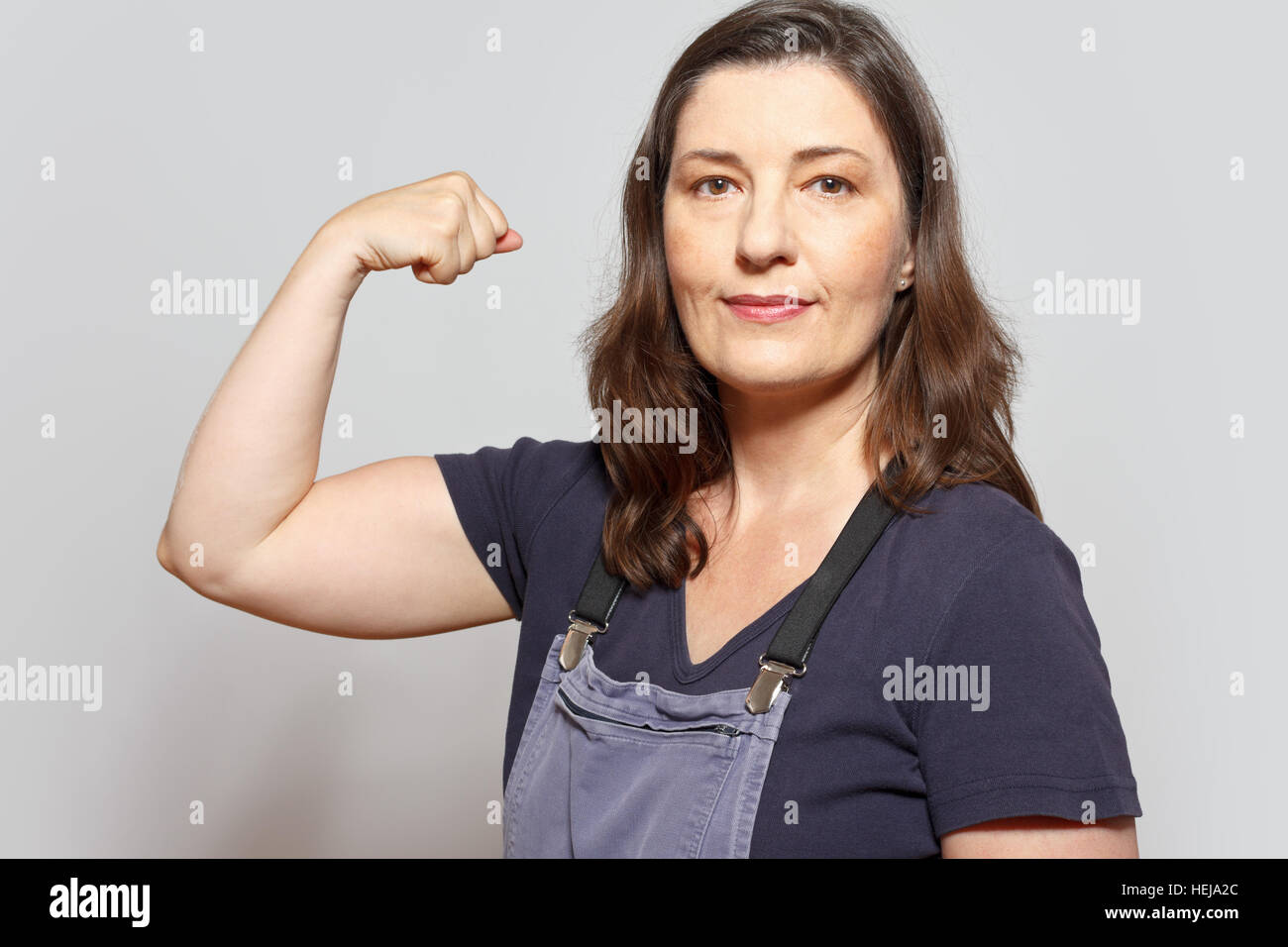Middle aged and self-confident woman in dungarees flexing her biceps muscles, sign for assertiveness, light gray background Stock Photo