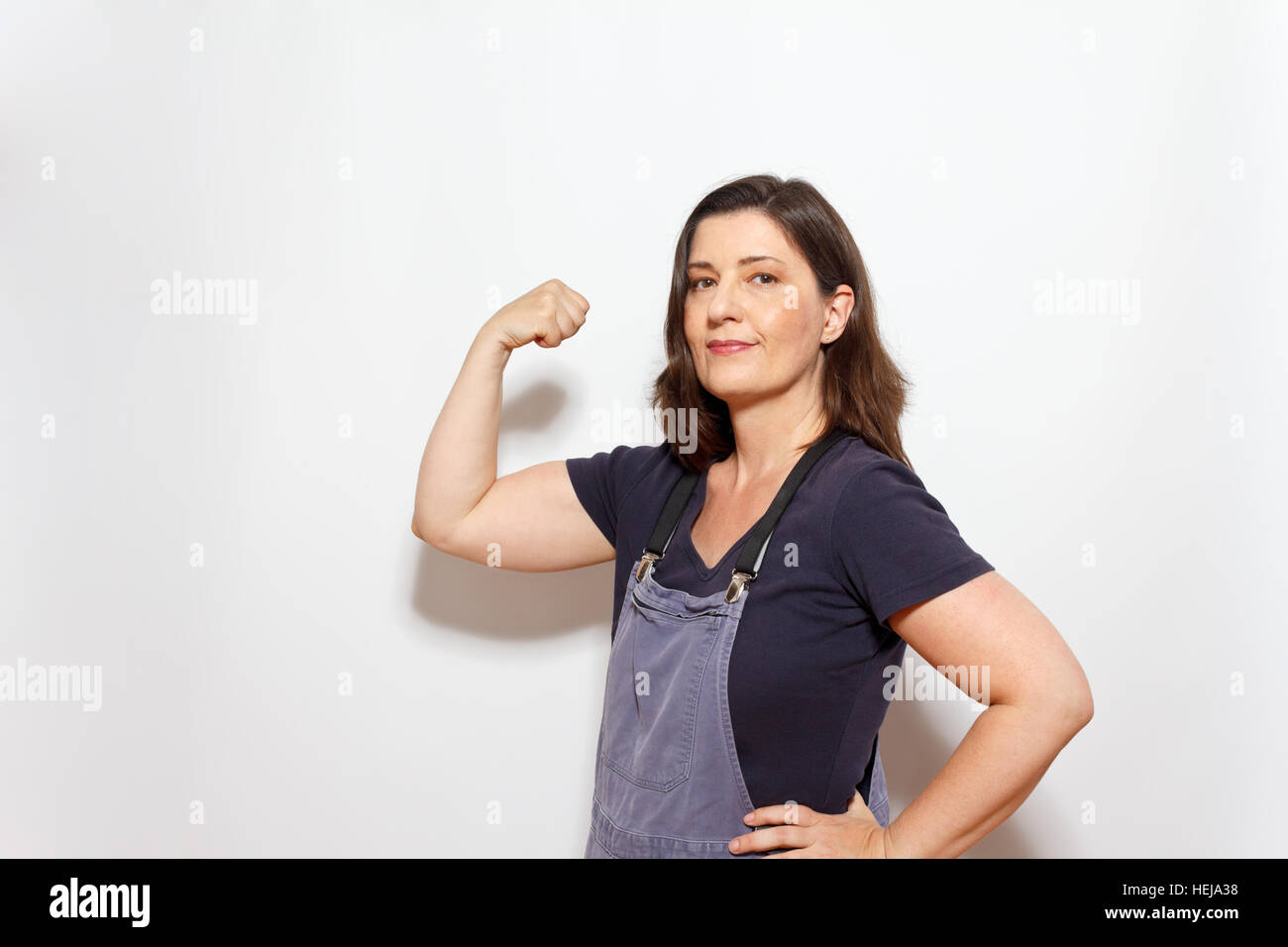 Middle aged woman in dungarees with hand on her hip and flexing the biceps muscles of the other arm, white background,copy space Stock Photo