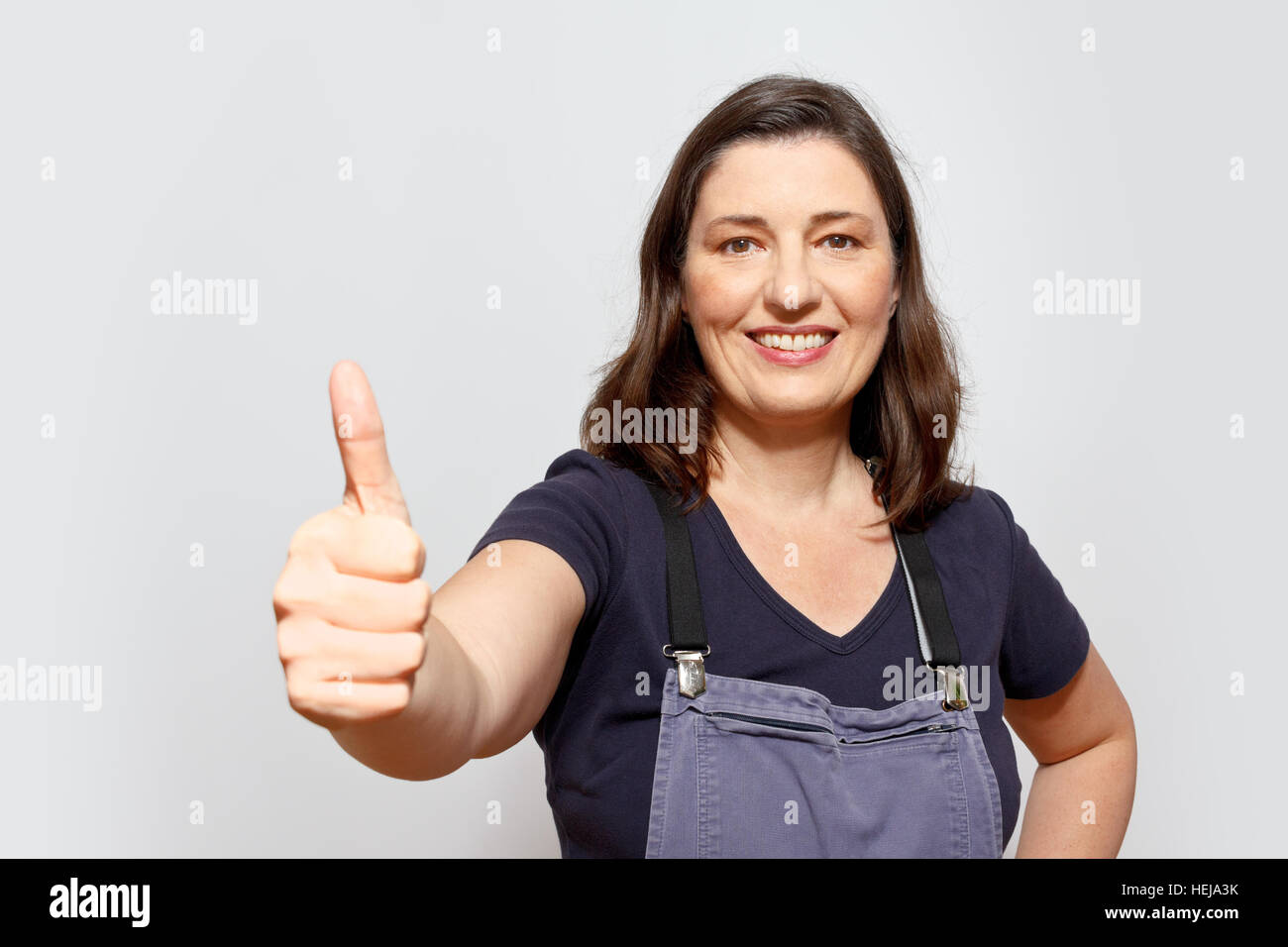 Smiling adult woman wearing dungarees showing the thumbs up sign, light grey or gray background, copy or text space Stock Photo