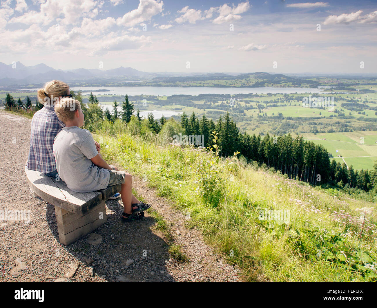 Mutter und Sohn sitzen auf einer Bank und blicken vom Berg ins Tal über das Ost-Allgäu, die Voralpen und Alpen Stock Photo