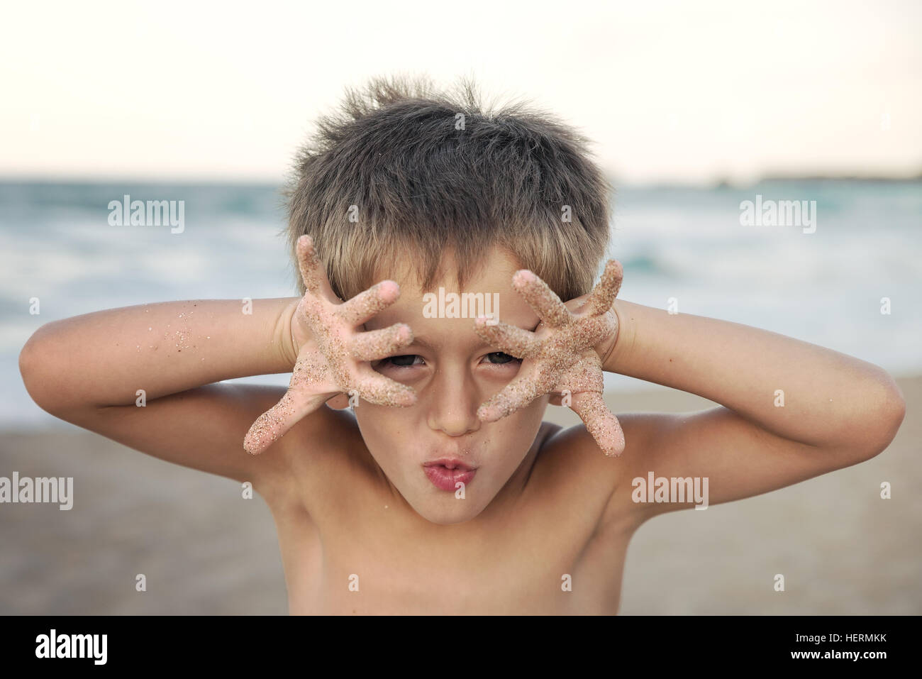 Boy standing on beach messing about with hands in front of face Stock Photo