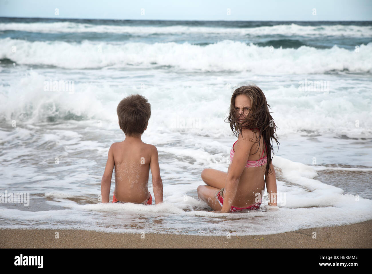 Girl and boy sitting on beach in water's edge Stock Photo