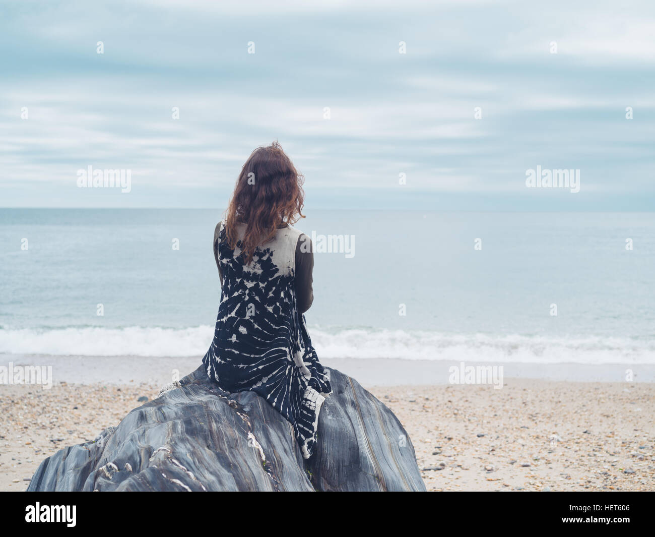 A young woman is sitting on a rock .on the beach and is looking at the ...