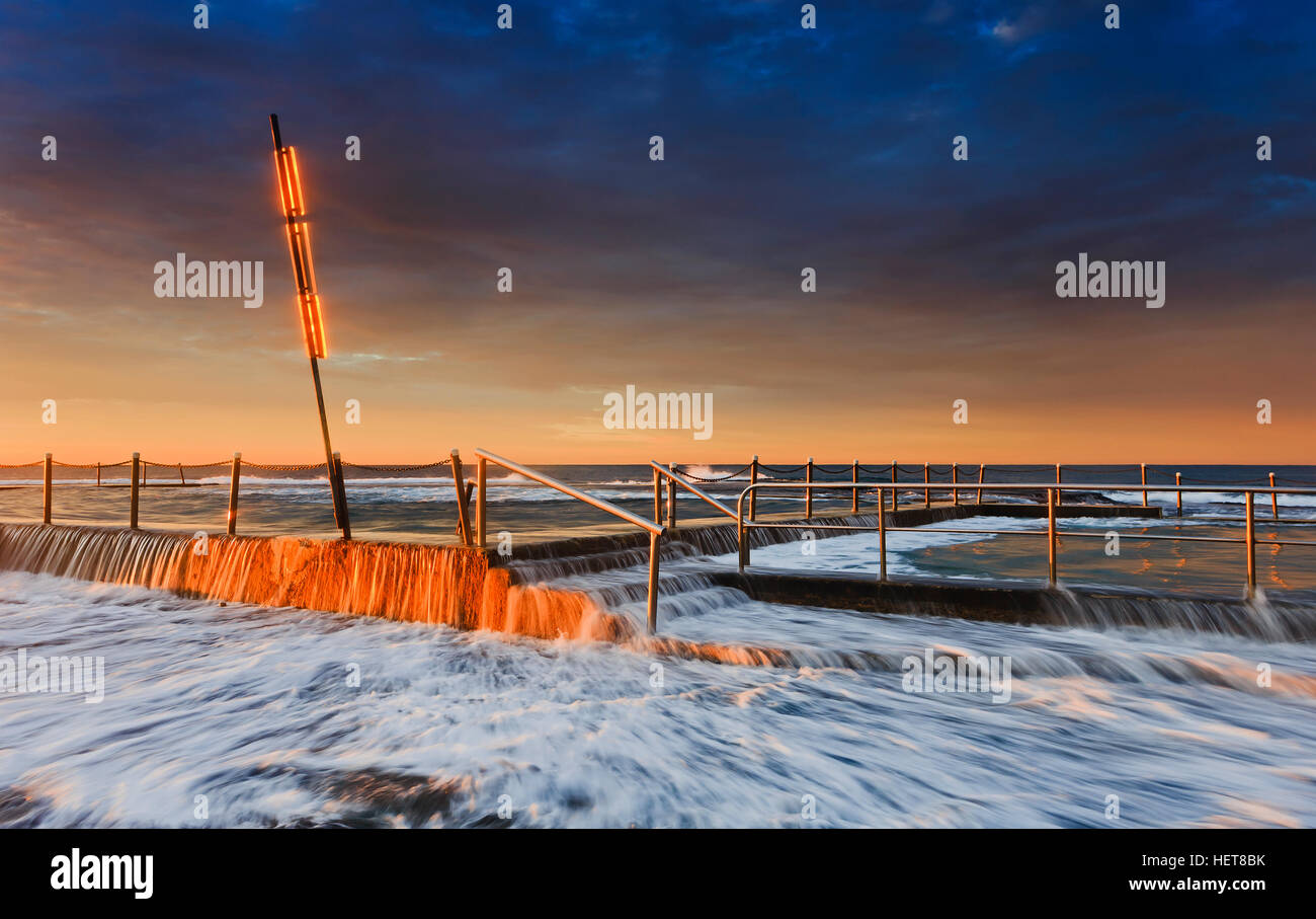 High rise surfing waver flowing over the rock pool at Mona Vale beach in Sydney. Pool fence and sign pole in warm rising sun light above surf foam. Stock Photo