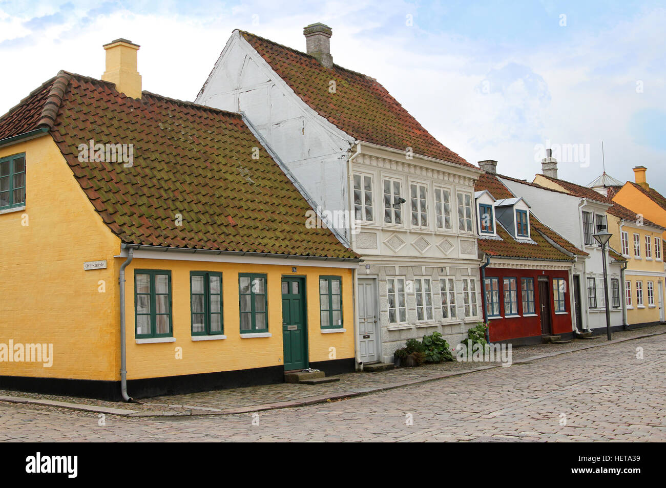A charming cobblestone street (Overgade) in Odense, Denmark, lined with colorful traditional Danish buildings featuring unique architectural details and historic lanterns.