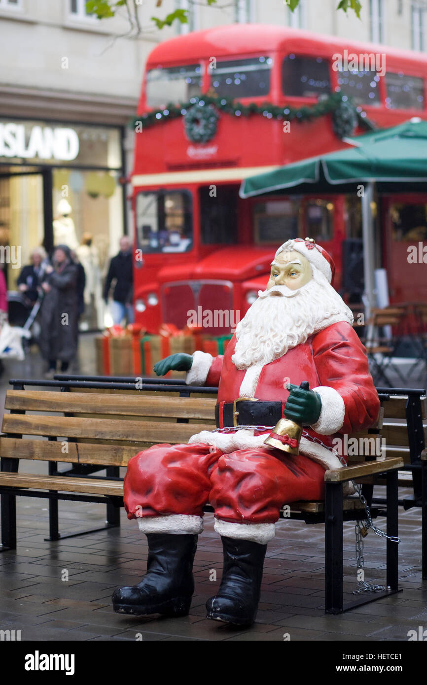 Santa Claus statue sitting on a bench with a red double decker bus in