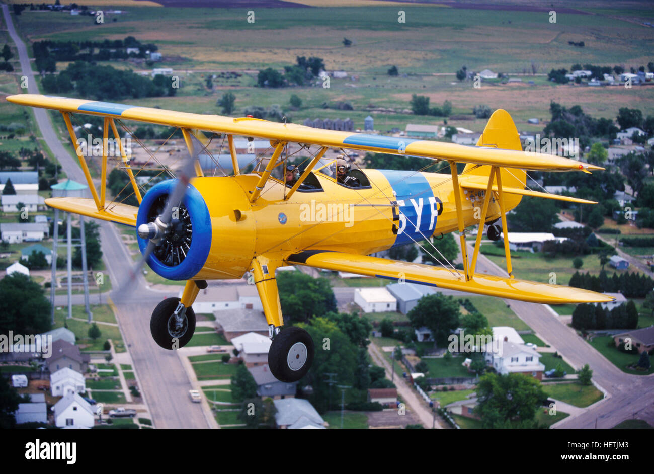 A yellow open cockpit Stearman biplane flies low over a small midwestern town. Stock Photo