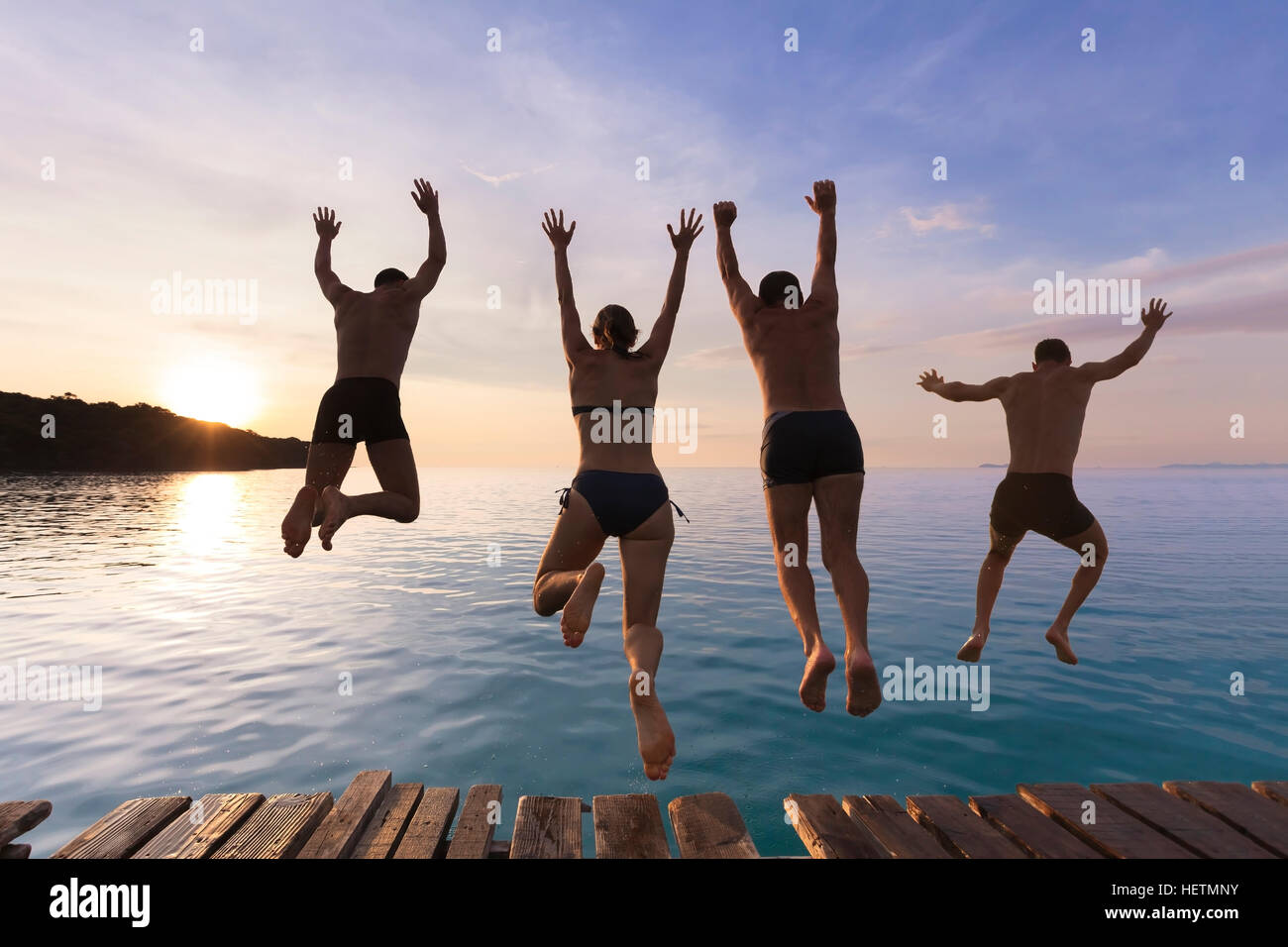 Cheerful people having fun jumping in the sea water from a pier at sunset Stock Photo