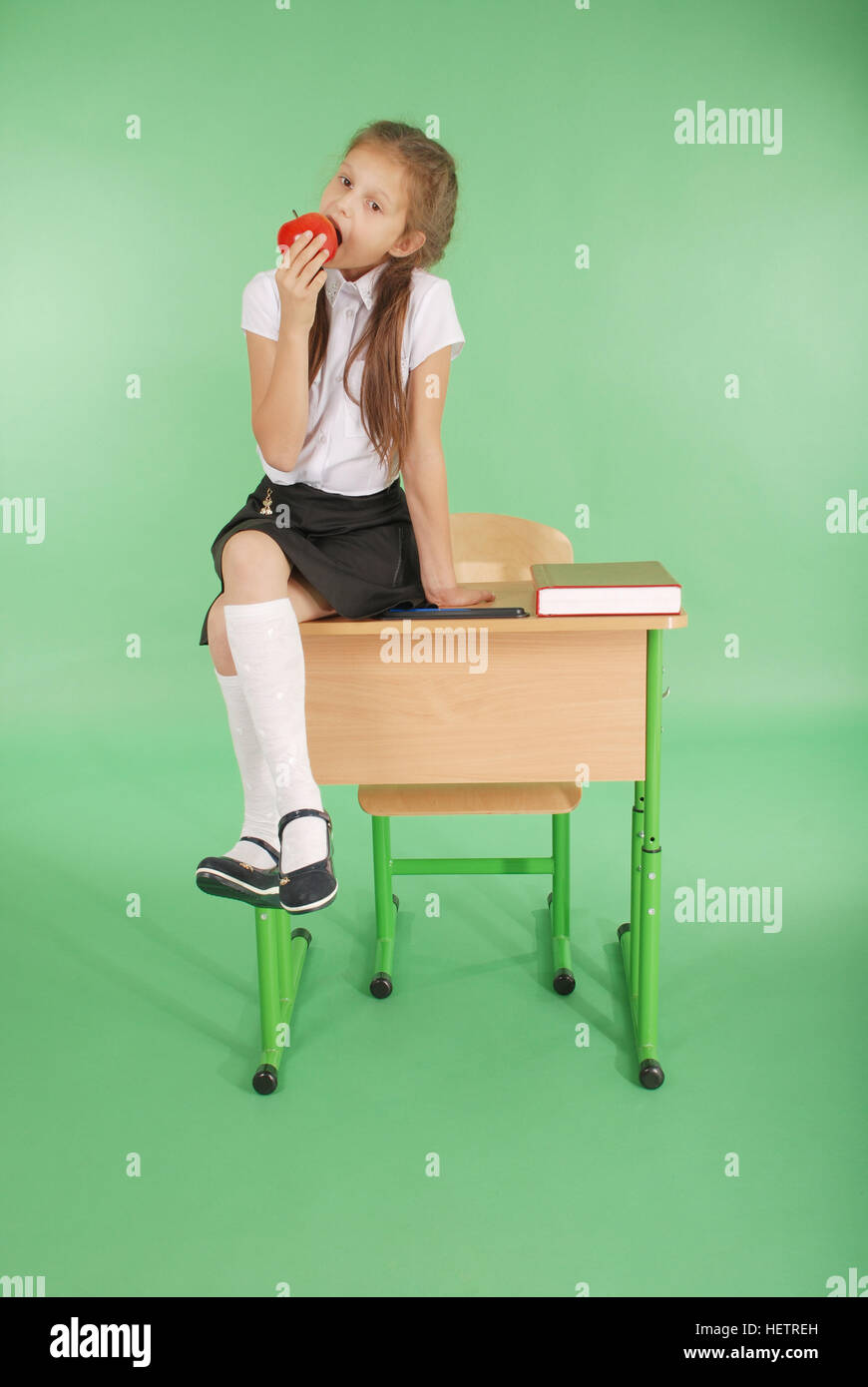 Girl in a school uniform sitting on desk and eat apple isolated on ...
