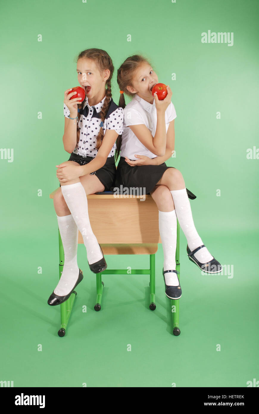 Two girls in a school uniform sitting on desk and eat apples isolated on green Stock Photo