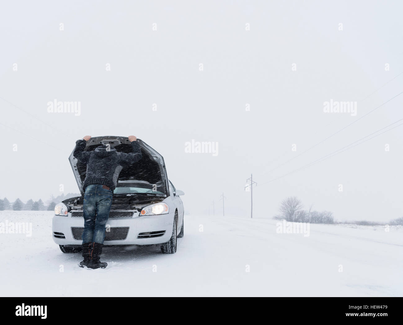 Rear view of man looking under hood of broken down car on snow covered landscape Stock Photo