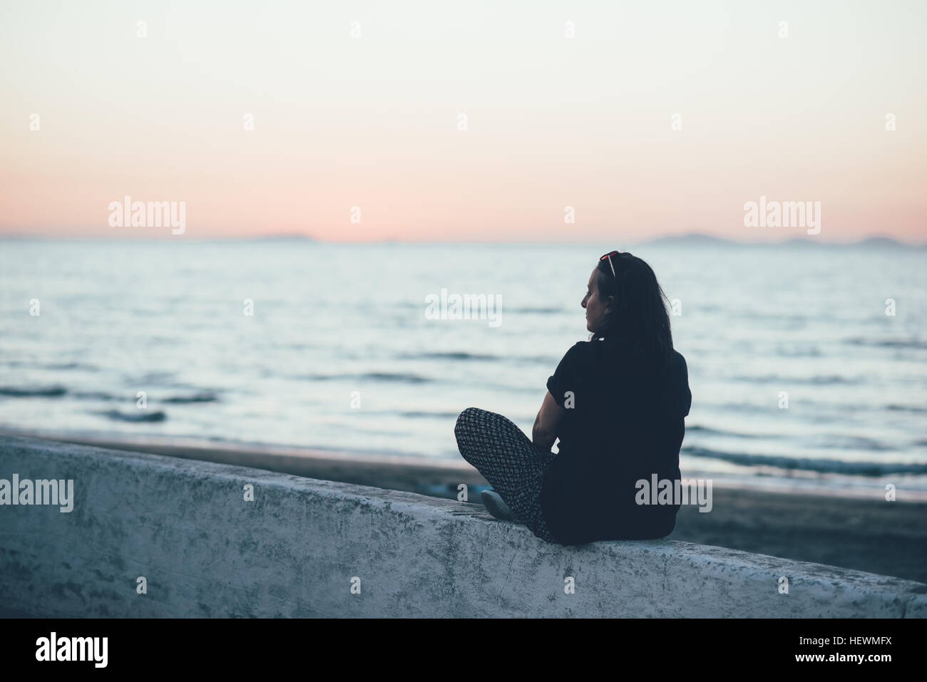 Rear view of woman sitting on wall looking at ocean, Sorso, Sassari ...
