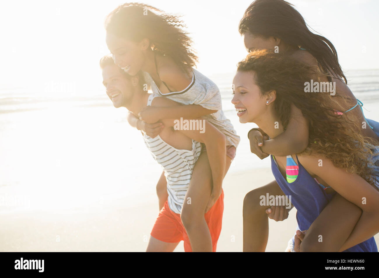 Young man and woman piggybacking friends on sunlit beach, Cape Town, South Africa Stock Photo