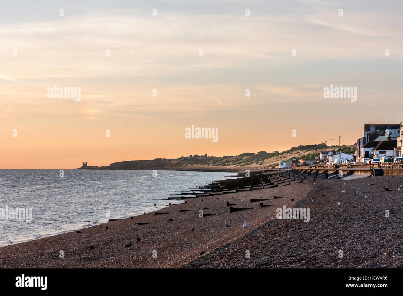 View along shingle beach with rows of wooden wave breakers, seafront promenade and town during Dawn. In distance, headland at Reculver. Stock Photo