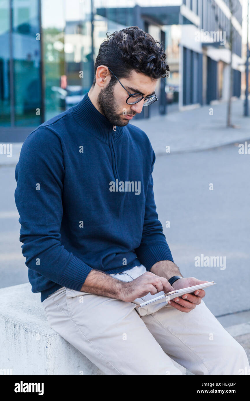 Young man sitting on bollard using mini tablet Stock Photo
