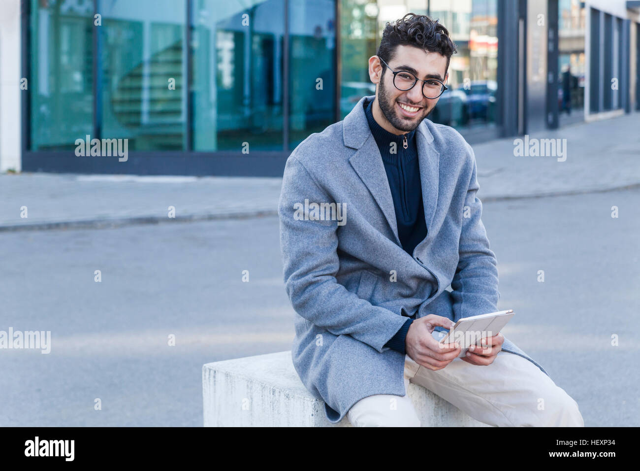 Portrait of smiling young man with mini tablet sitting on bollard Stock Photo
