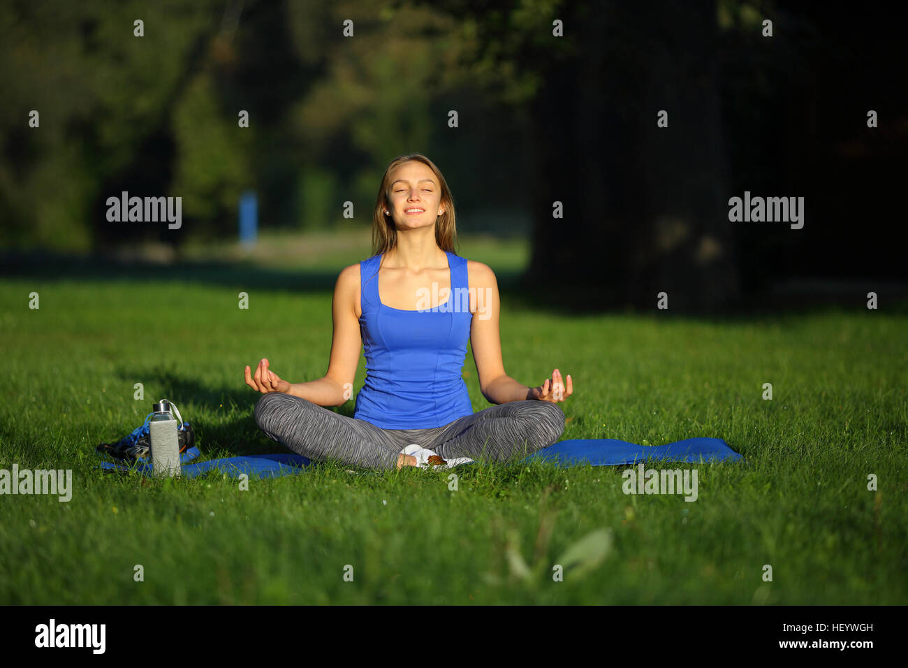 Smilling young woman sitting in lotus position on yoga mat in park ...