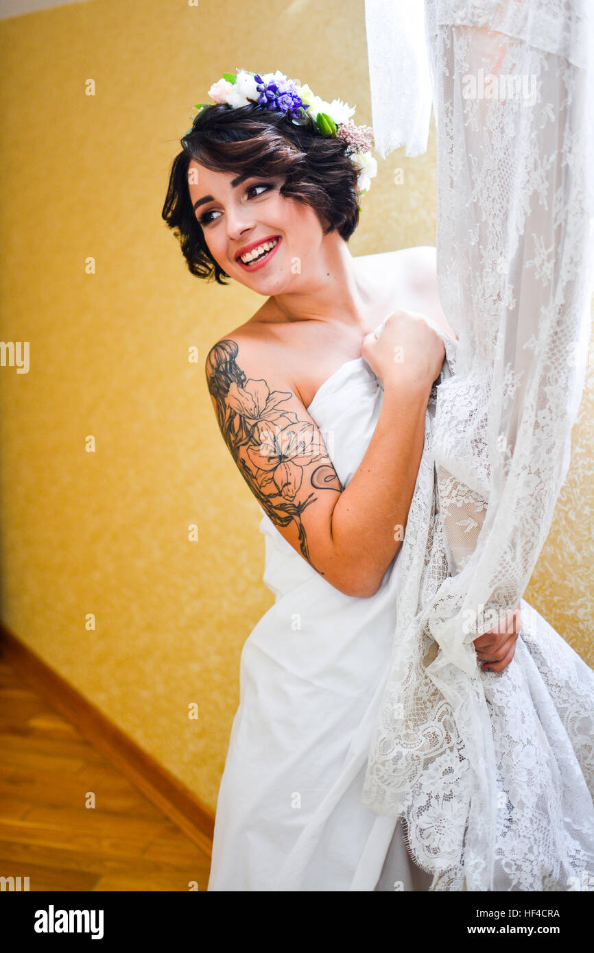 Beautiful smiling brunette bride posing with his wedding lace dress in hand and looking at mirror. Emotional girl wearing white  flowers in hair. Stock Photo