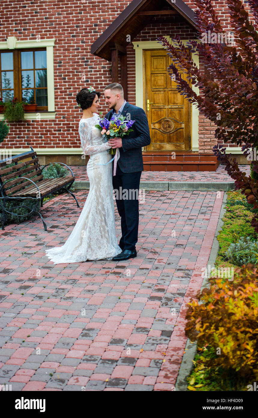 Portrait of emotional newlyweds groom and bride embracing  holding each other hands. In yard against house in autumn season. Stock Photo
