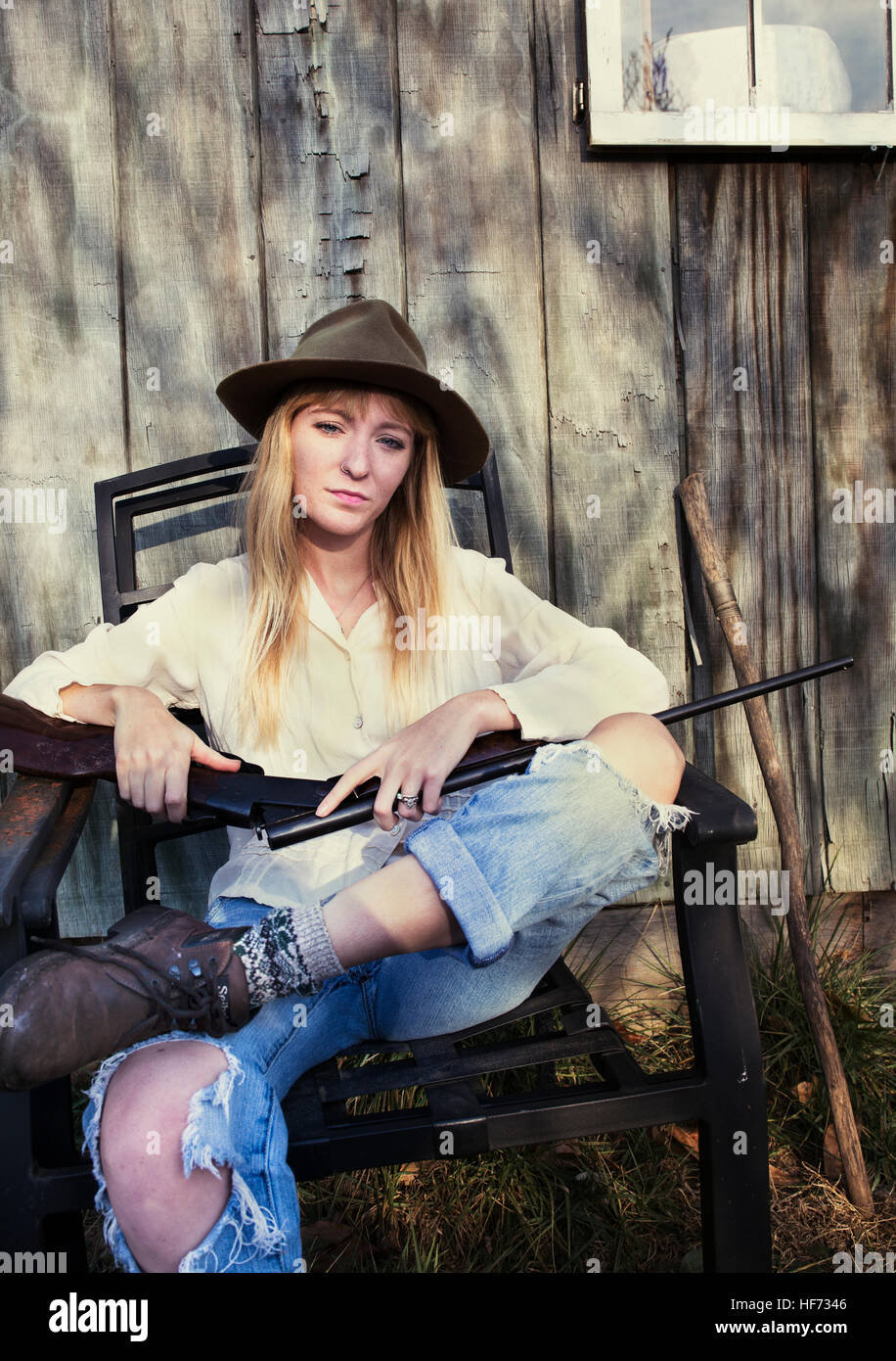Beautiful blond woman holding rifle sitting in front of a wooden wall Stock  Photo