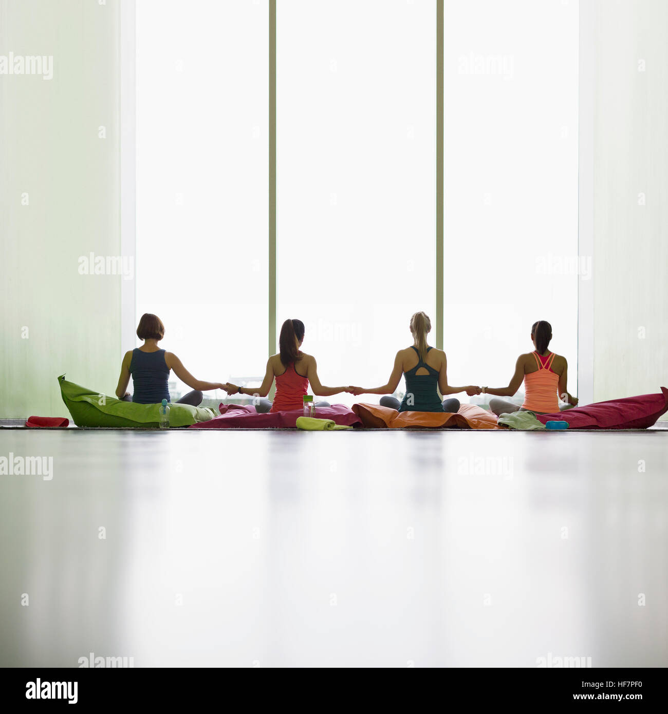 Women on cushions holding hands at window in restorative yoga gym studio Stock Photo