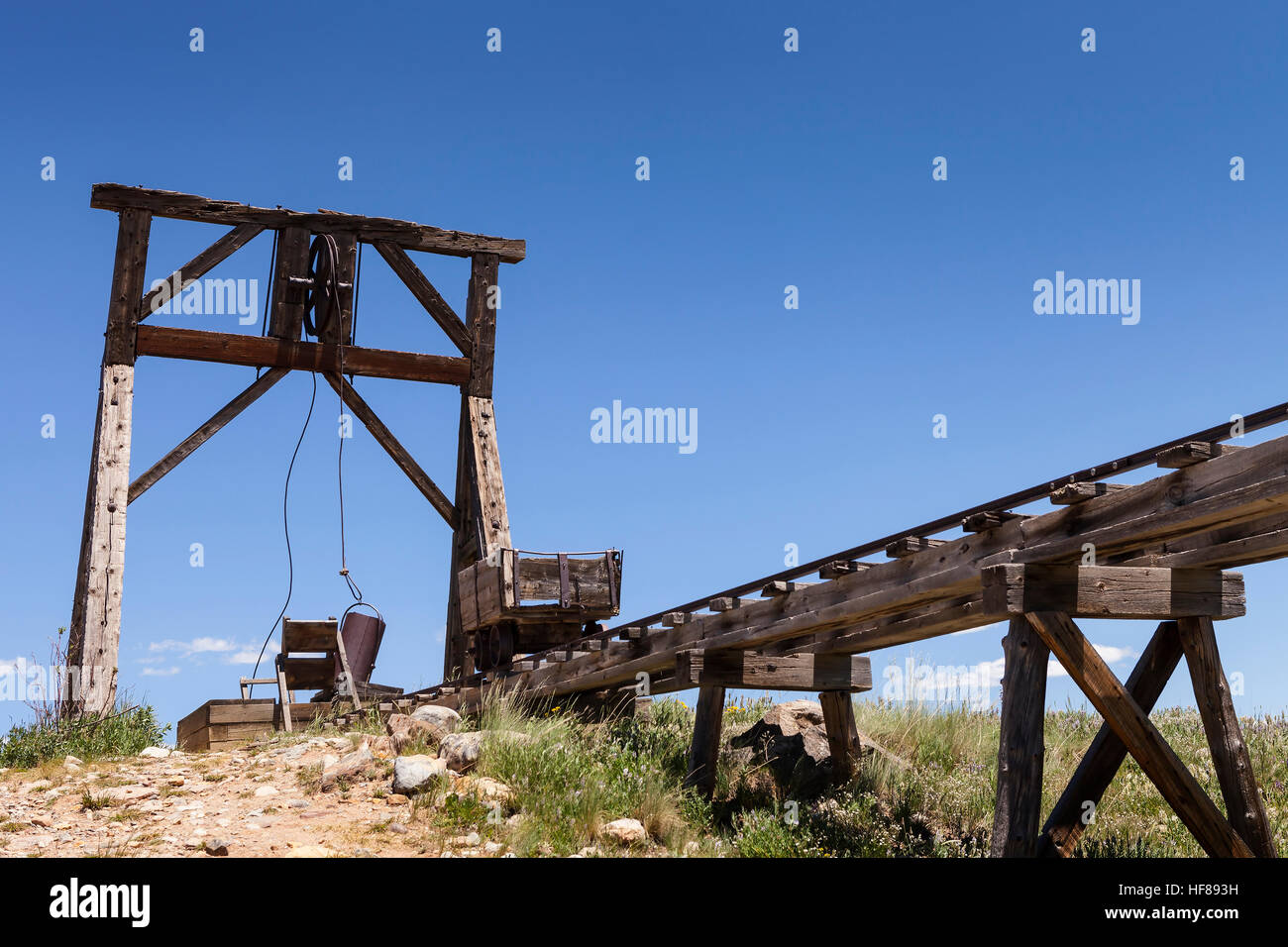 Old mining ore cart on trestle with head frame under blue sky. Stock Photo