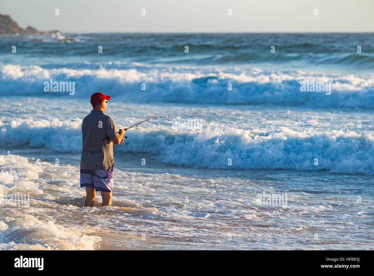 An older man standing on a beach fishing in the surf with a beach fishing rod at a Sydney beach early on a summer morning in Australia Stock Photo