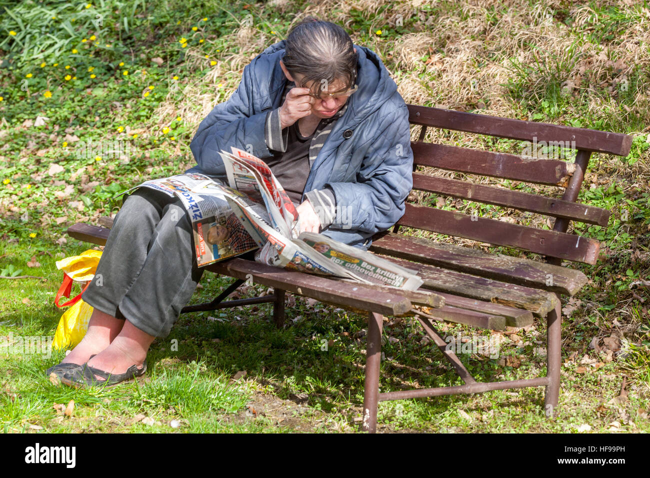 An elderly senior woman reading newspapers in a city park on a bench, failing eyesight Stock Photo