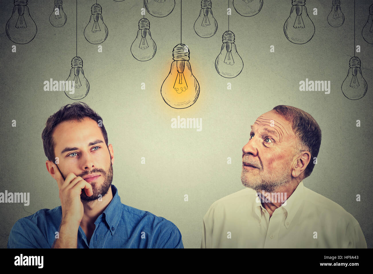 Cognitive skills concept, old man vs young person. Senior man and young guy looking at bright light bulb isolated on gray wall background Stock Photo