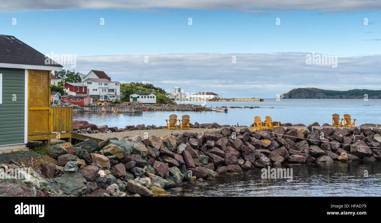 Golden adirondack chairs on a rock jetty.   Houses on the sea along a village shoreline.  Rural Newfoundland, Canada. Stock Photo