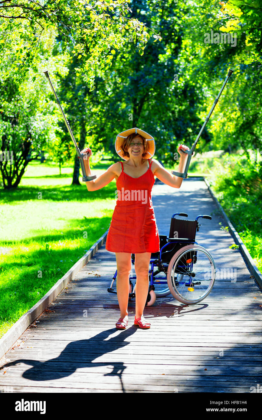 handicapped woman standing ad raising her crutches in the park, wheelchair in the background Stock Photo