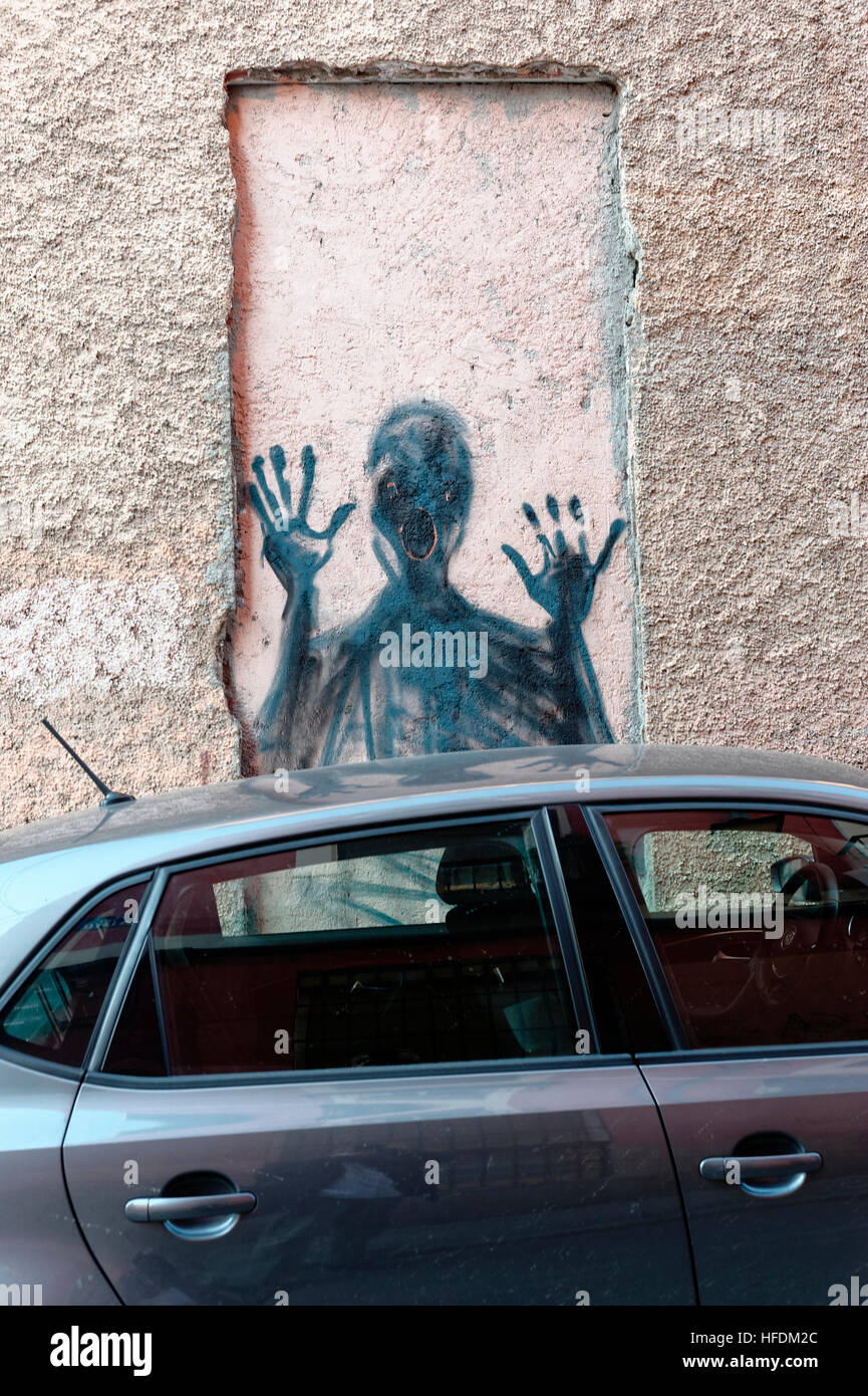 Clever disturbing street art of a person screaming through a window in a wall, over the top of a parked car on a Madrid wall, Stock Photo