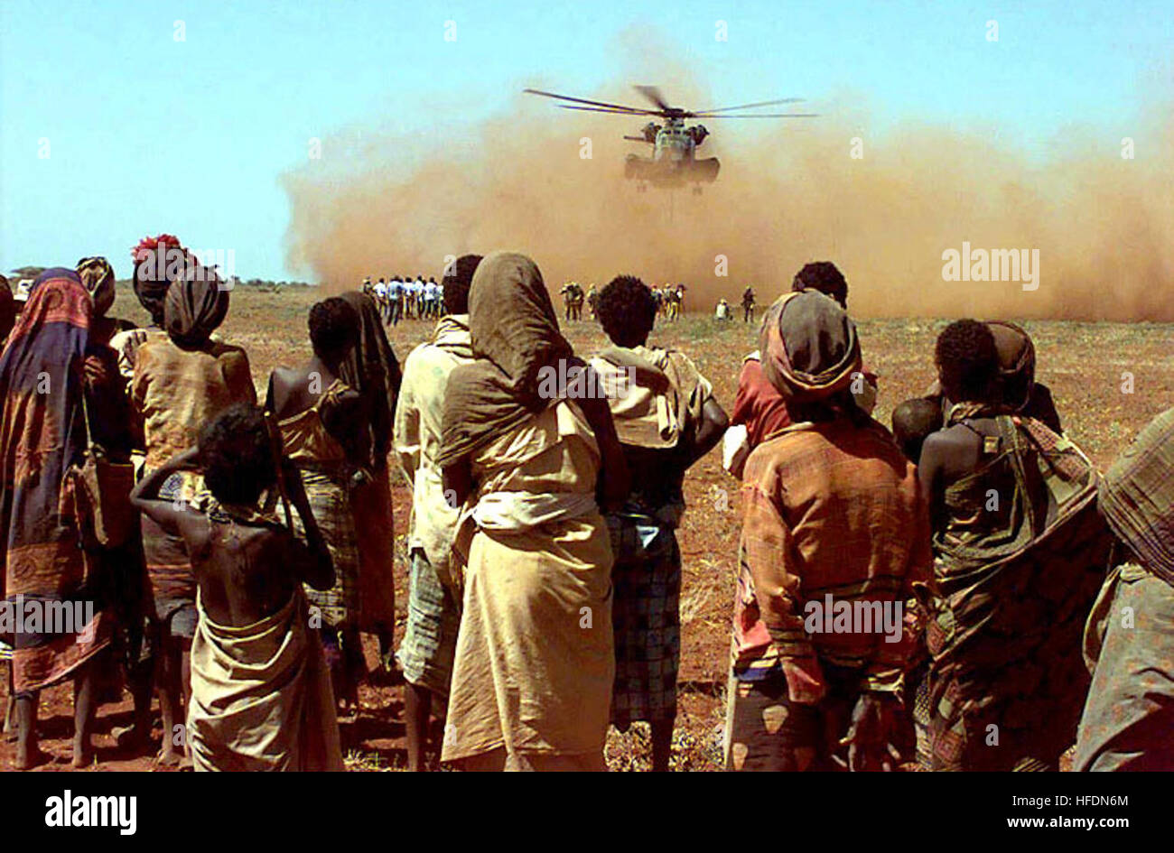 The camera shoots past some Somalis from the village of Maleel as they watch a US Marine CH-53 Sea Stallion deliver a sling load of wheat donated by the people of Australia.  Several Somali men and Australian Army Soldiers wait near the loading zone where the helicopter rotor wash is kicking up large amounts of red dirt.  This mission is in direct support of Operation Restore Hope. Aus wheat in Somalia Stock Photo