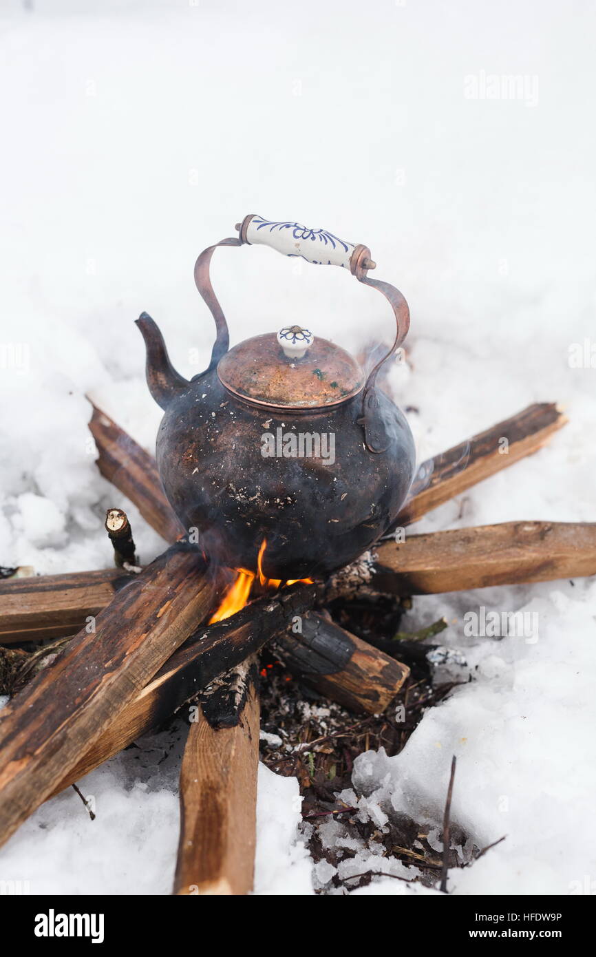 Copper kettle over an open fire in winter. Boiling kettle on firewood. Open fire cooking. Snow around. Lifestyle, camping. Blurred background. Stock Photo