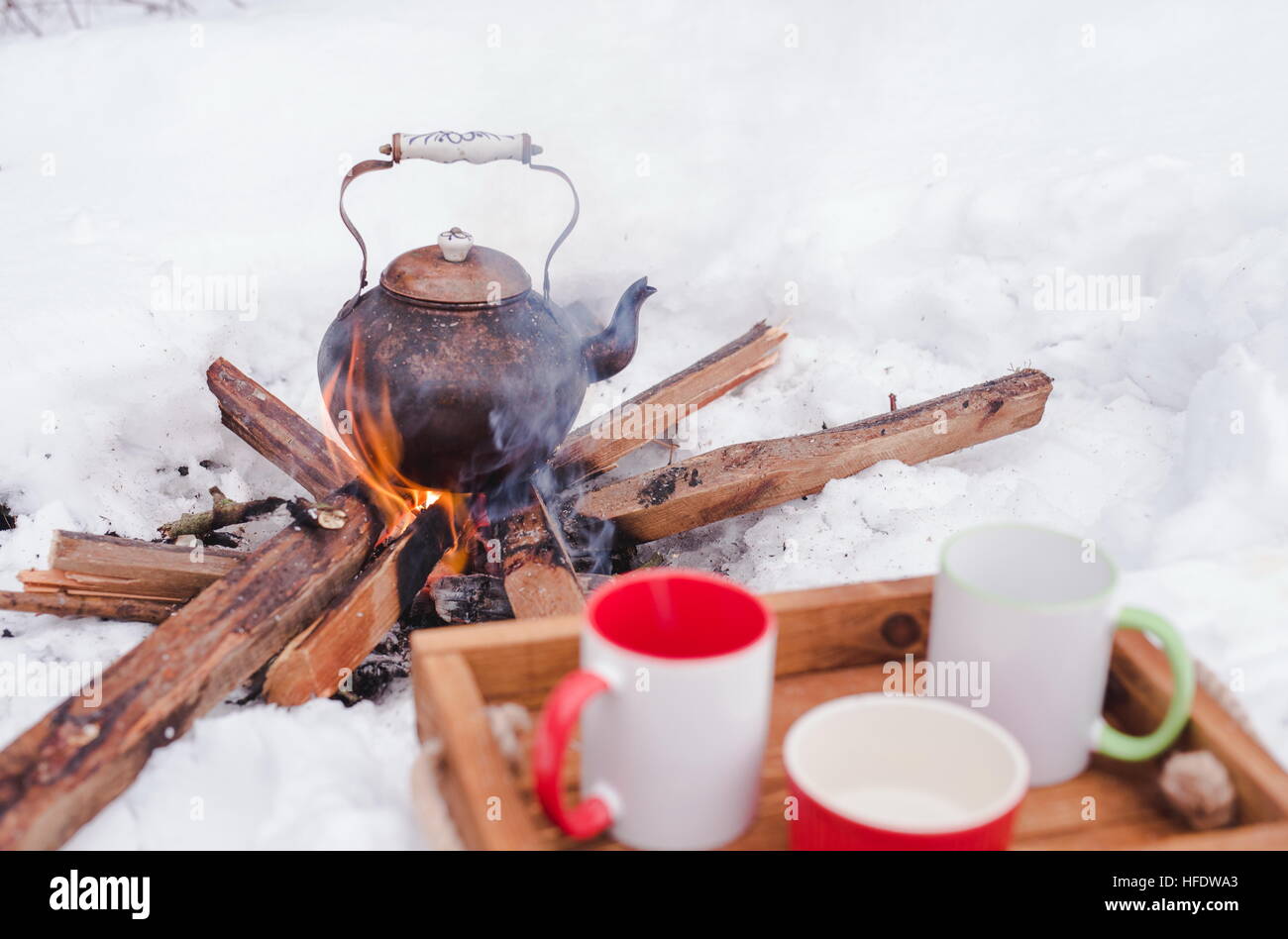 Romantic winter picnic. Two cups and a bowl on a wooden tray in snow. Copper kettle over an open fire on background, blurred. Boiling kettle on firewo Stock Photo