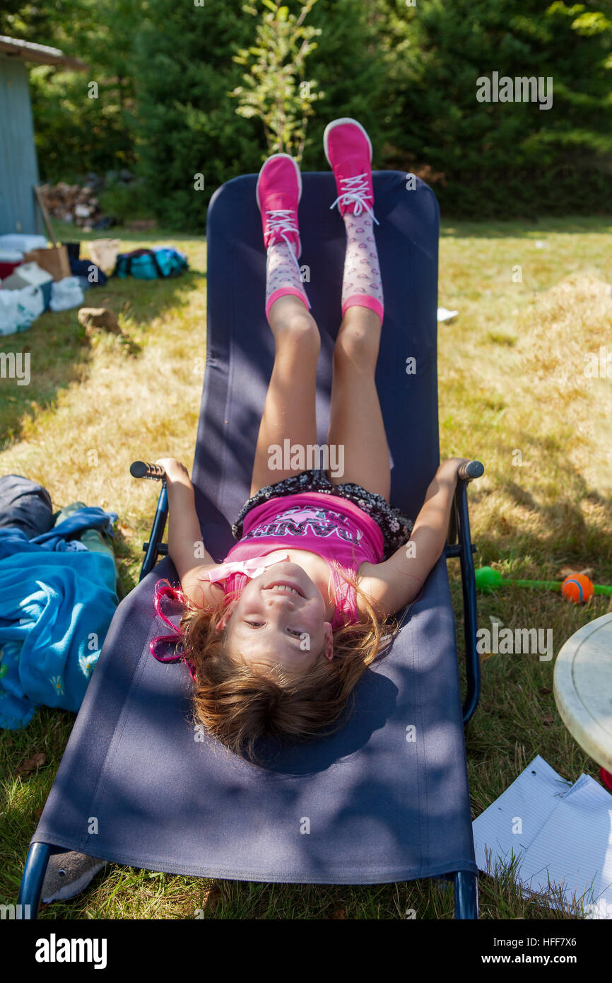 A cute seven-year old girl lays upside down on a blue chaise lounge at a camp in Vermont, United States. Stock Photo