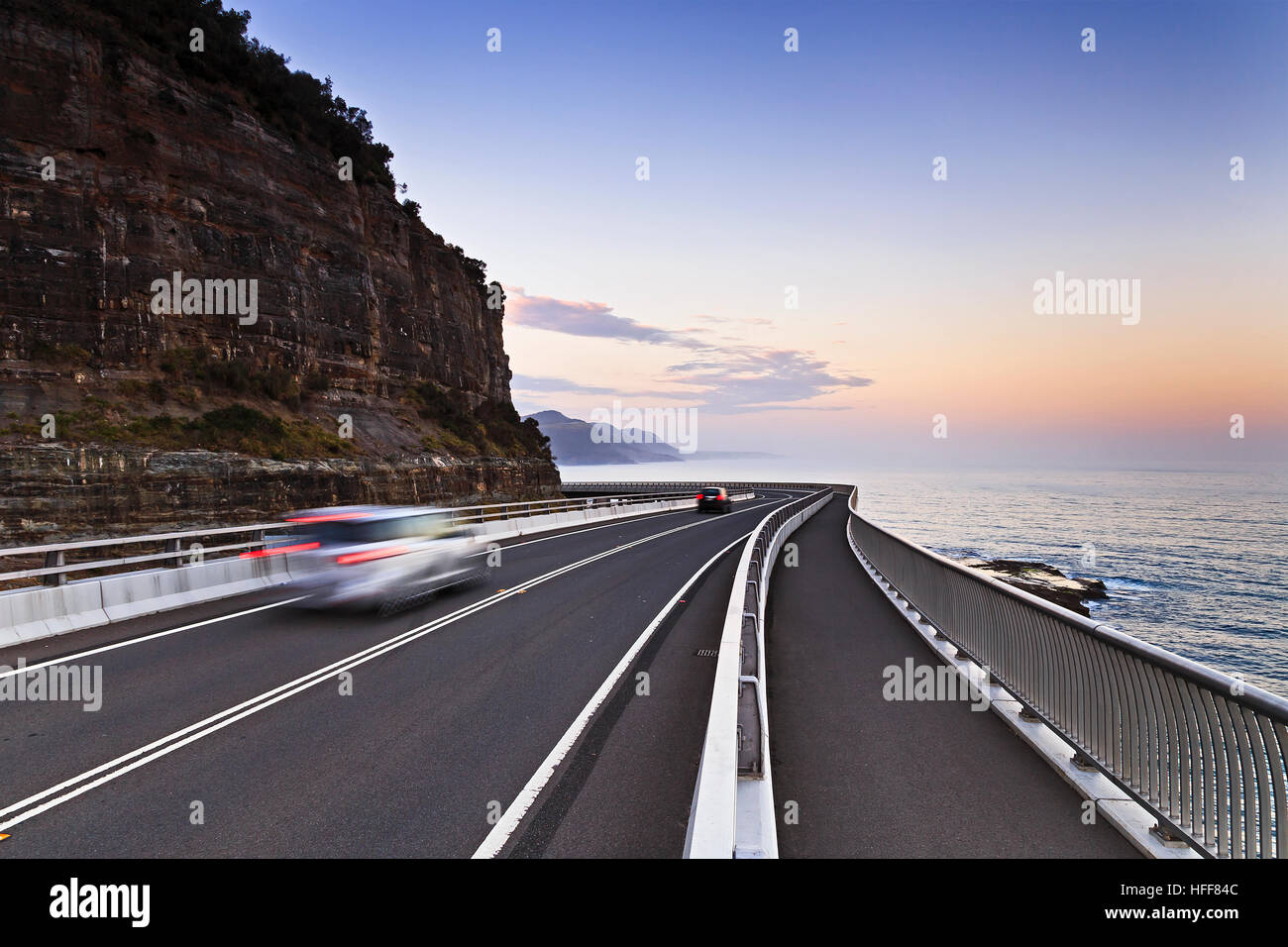Sea Cliff bridge of Grand Pacific Drive along the NSW coast of Australia on Pacific at sunset. Scenic tourist drive with blurred cars around cliff roa Stock Photo