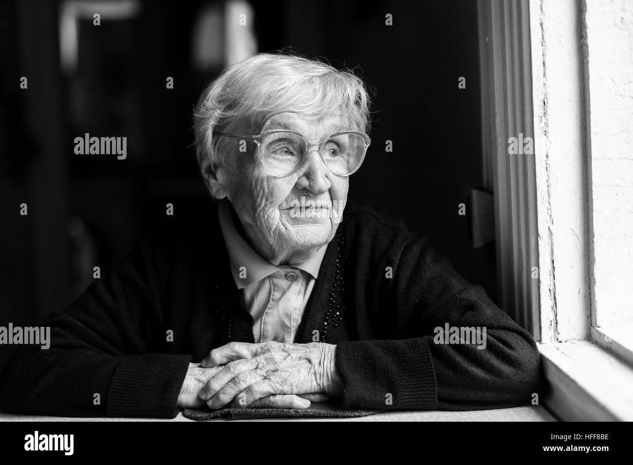 An elderly woman with glasses sitting at a table, a black-and-white photo. Stock Photo