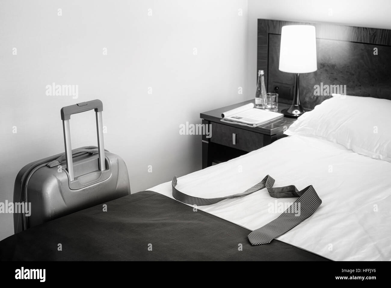 Black and white picture of a tie on bed and suitcase in empty hotel room, conceptual picture. Stock Photo
