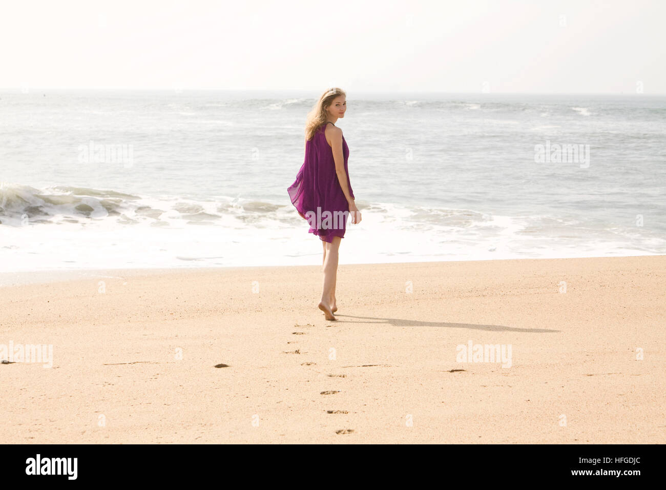 Young woman walking towards the water on a beach leaving footprints in the sand while looking over her shoulder Stock Photo
