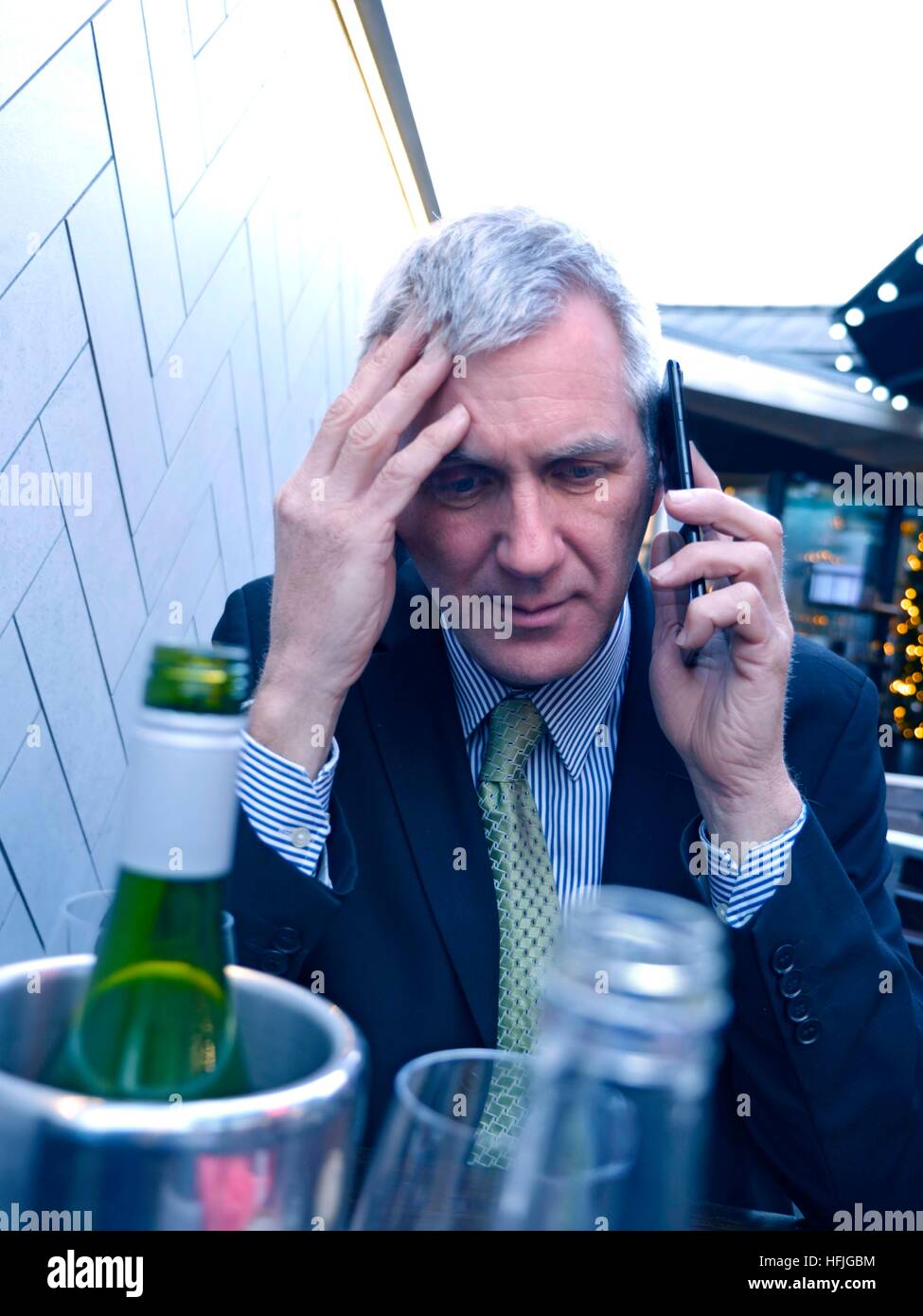 Businessman with alcoholic drinks on table looking worried, bad news, pensive, concerned, listening on his smartphone  iPhone 7 plus mobile telephone at alfresco drinks  bar restaurant table with alcohol wine bottles and glass in foreground Stock Photo