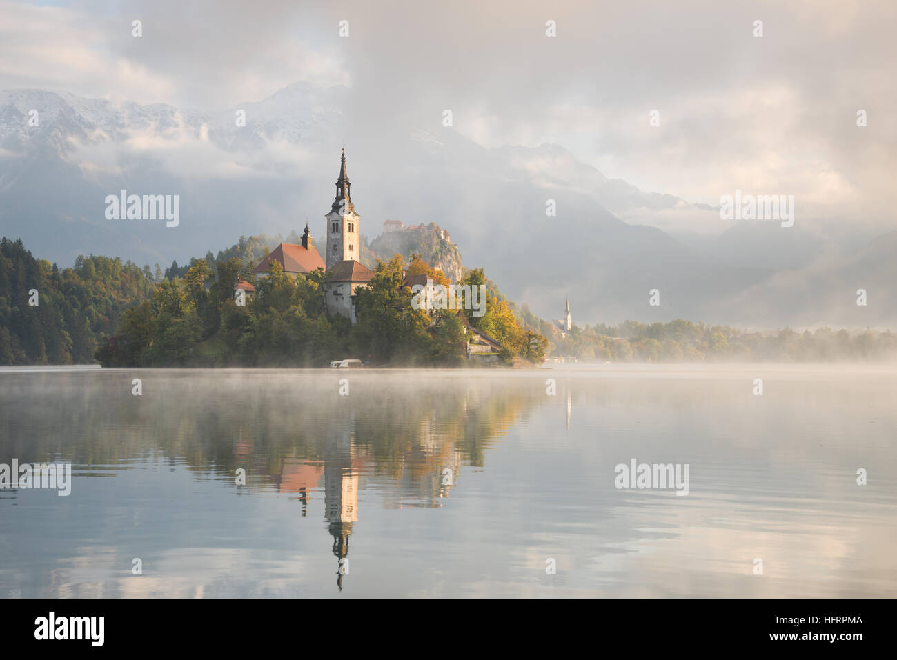 Church on the small island in Lake Bled at sunrise on a foggy morning in autumn with reflection in the water in Slovenia. Stock Photo