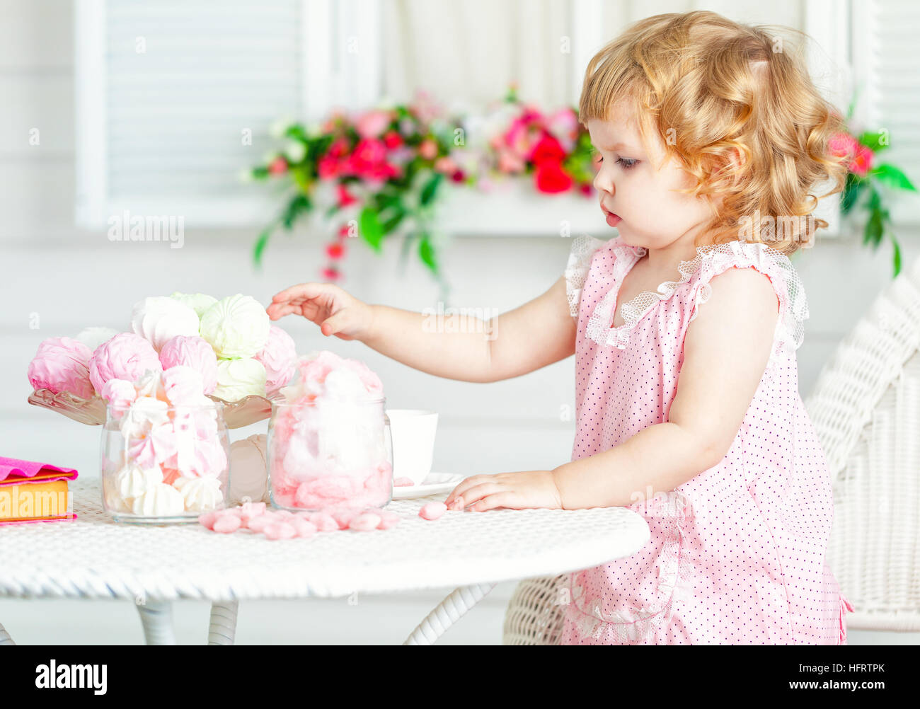 Little cute curly girl in a pink dress with lace and polka dots sitting at the table in the garden and eating different sweets. Stock Photo