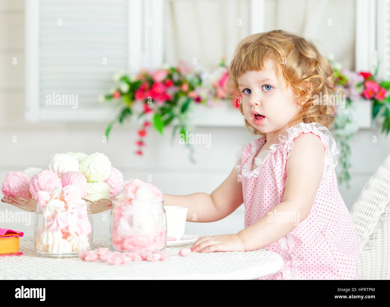 Little cute curly girl in a pink dress with lace and polka dots sitting at the table in the garden and eating different sweets. Stock Photo