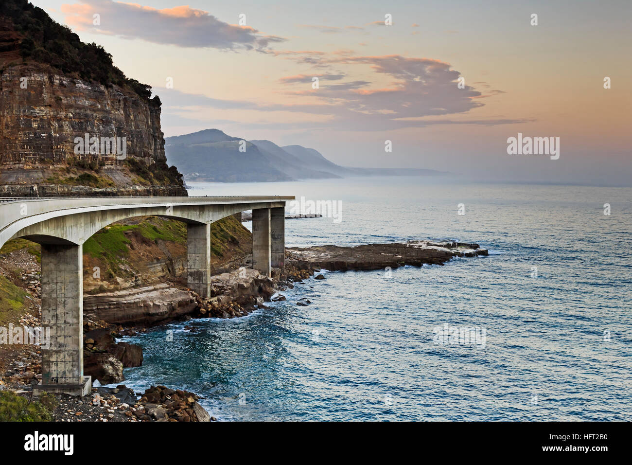 Side view of Sea Cliff Bridge on Grand Pacific Drive in Australia. Bright sunset over pacific ocean from scenic tourism motor way facing hilly coast. Stock Photo