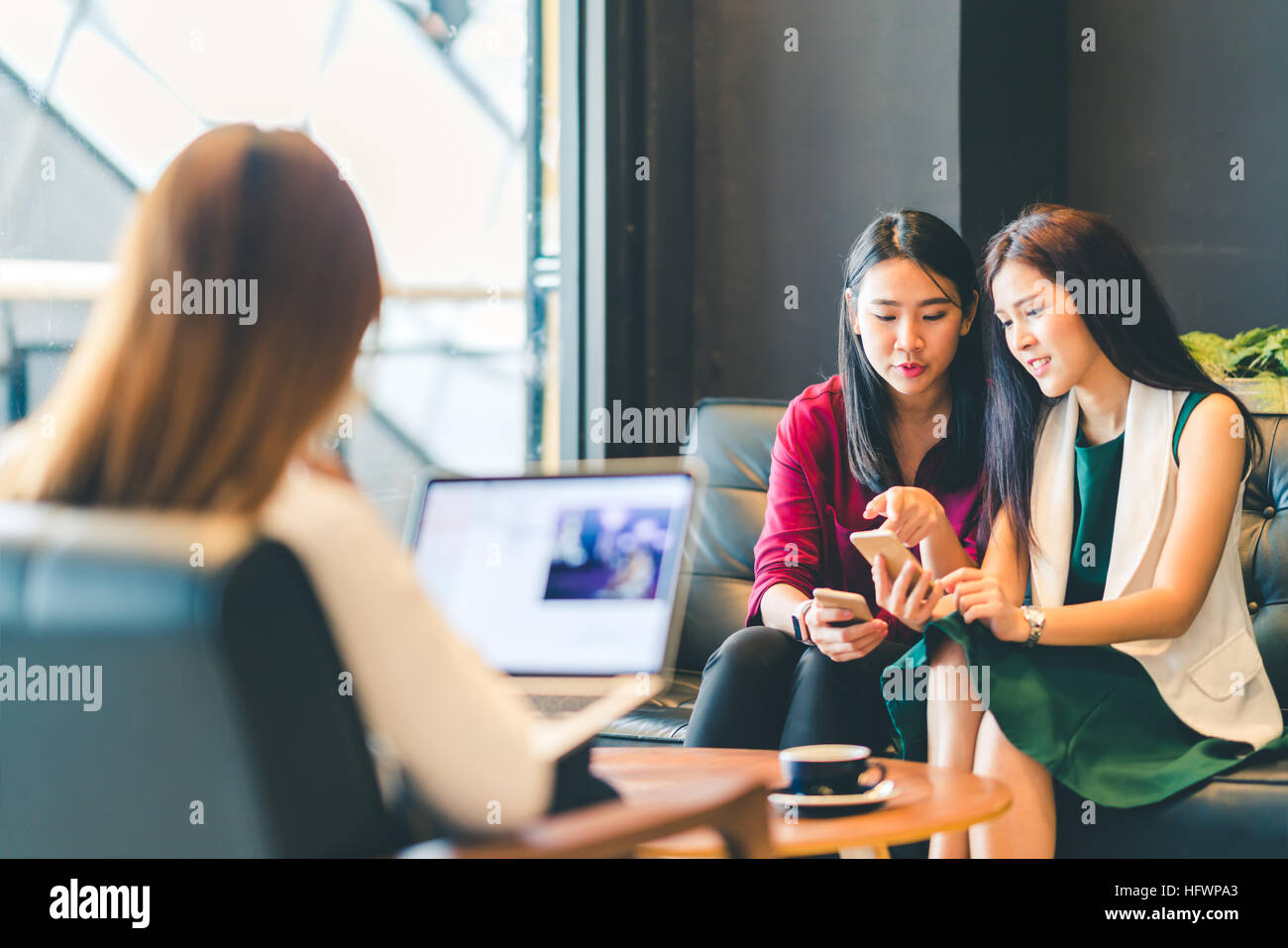 Beautiful Asian girls using smartphone and laptop, chatting on sofa at cafe, modern lifestyle with gadget technology or working woman on casual busine Stock Photo