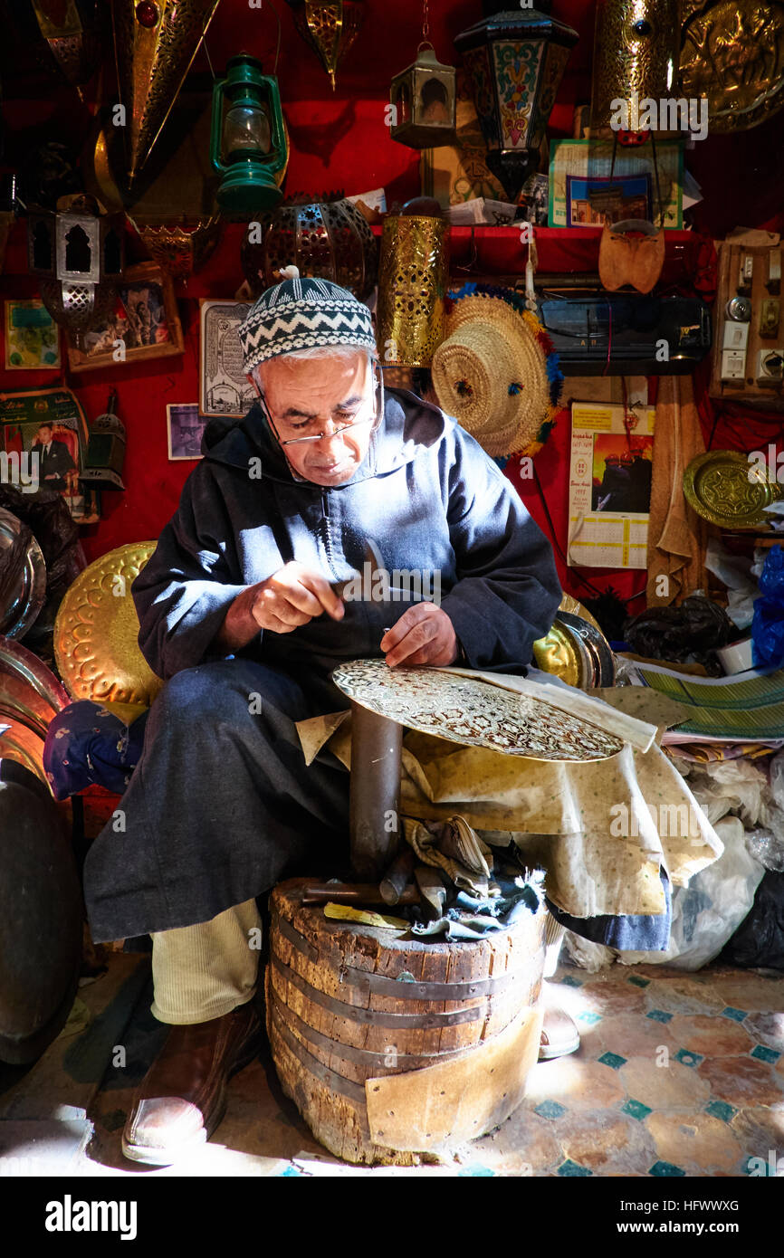 A Moroccan copper and silver-smith hammers plates in the Kettanine district of the Fez medina, Morocco surrounded by his work. Stock Photo