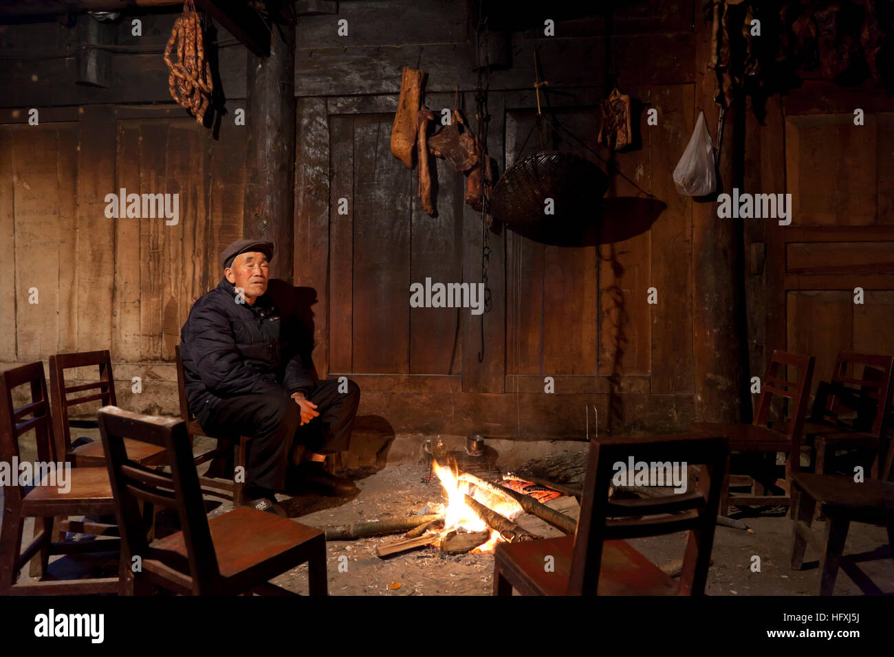 An old man in a village in the mountains of west China sits in the living room of his farmhouse near the fire to keep warm in winter. Stock Photo