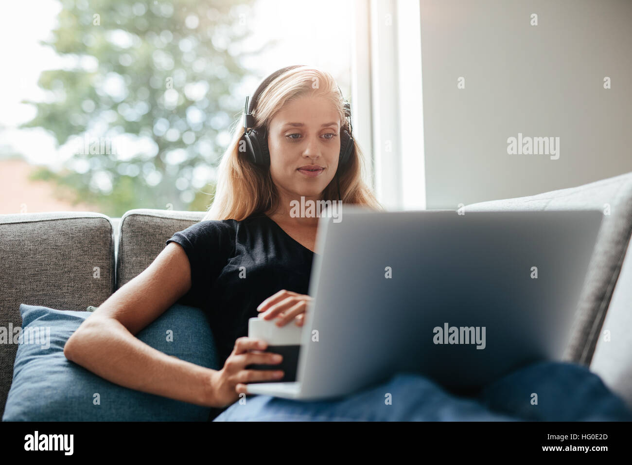 Young woman relaxing on sofa with laptop in living room. Female with headphones looking at laptop. Stock Photo