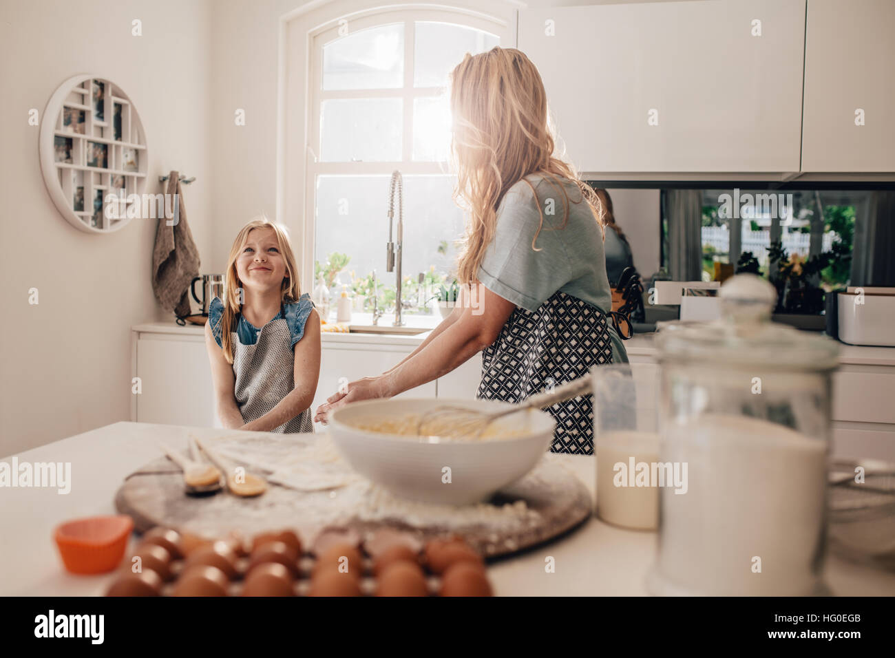 Happy little girl standing in kitchen and her mother cooking food. Mother and happy daughter baking in kitchen. Stock Photo